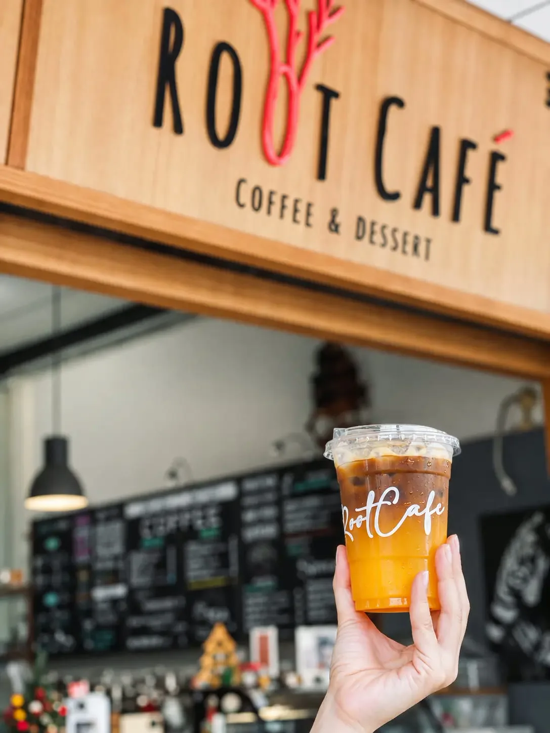 Iced coffee in a Root Cafe cup held up in front of the cafe's wooden sign