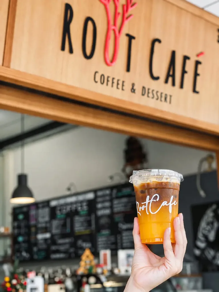 Iced coffee in a Root Cafe cup held up in front of the cafe's wooden sign